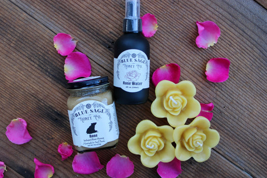 rose infused honey, rose water and floating rose beeswax candles on a rustic wooden surface surrounded with rose petals