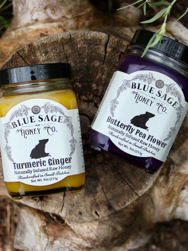 Two jars of Blue Sage Honey Co. honey, Turmeric Ginger and Butterfly Pea Flower, displayed on a rustic wood slice outdoors.